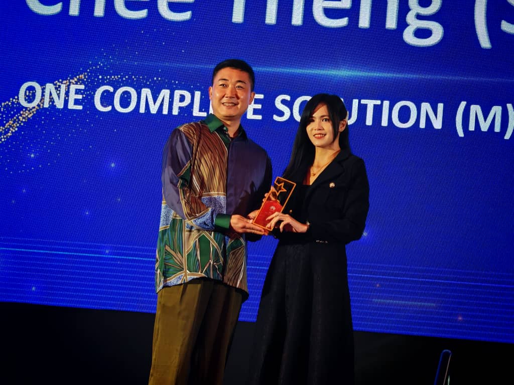A man and a woman stand on stage smiling, holding an award together. The background displays blue lighting and partially visible text. Both are dressed in formal attire.