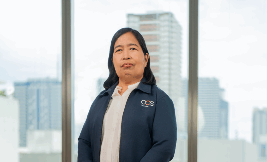 A woman with long dark hair, wearing a white blouse and a blue jacket with an OCS logo, stands indoors in front of large windows with a cityscape in the background.