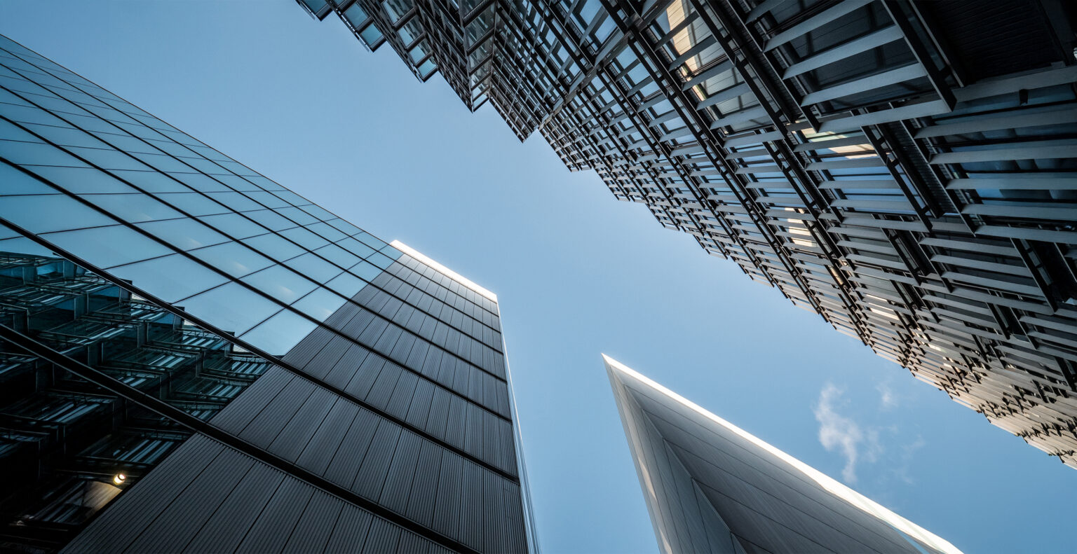 Looking up at modern skyscrapers with reflective glass and metal facades against a clear blue sky, creating geometric shapes and lines from a low-angle perspective.