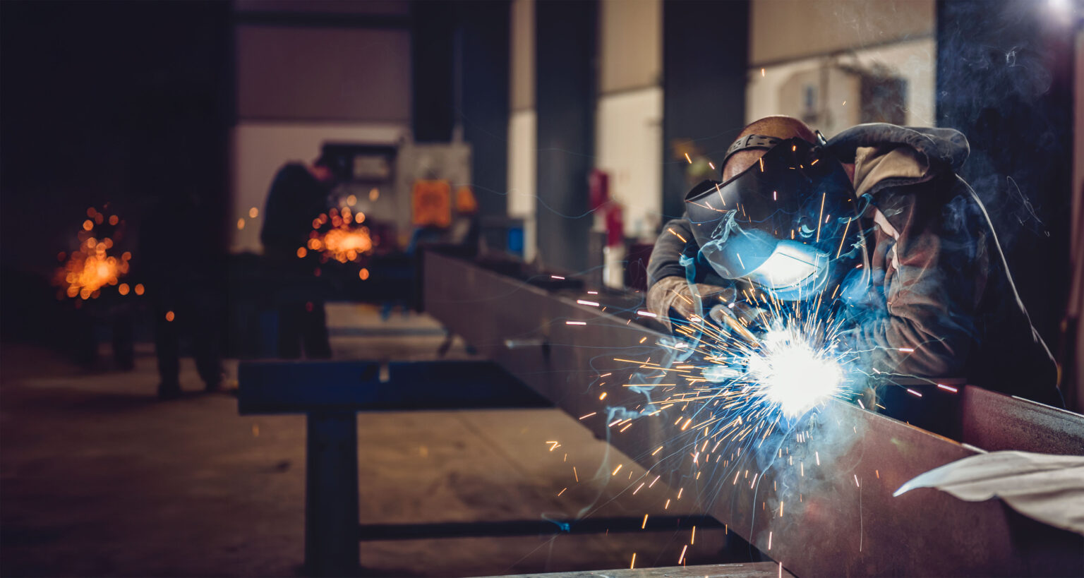 A welder in protective gear works on a metal beam, creating bright sparks in a workshop. Other workers and welding activities are visible in the background.