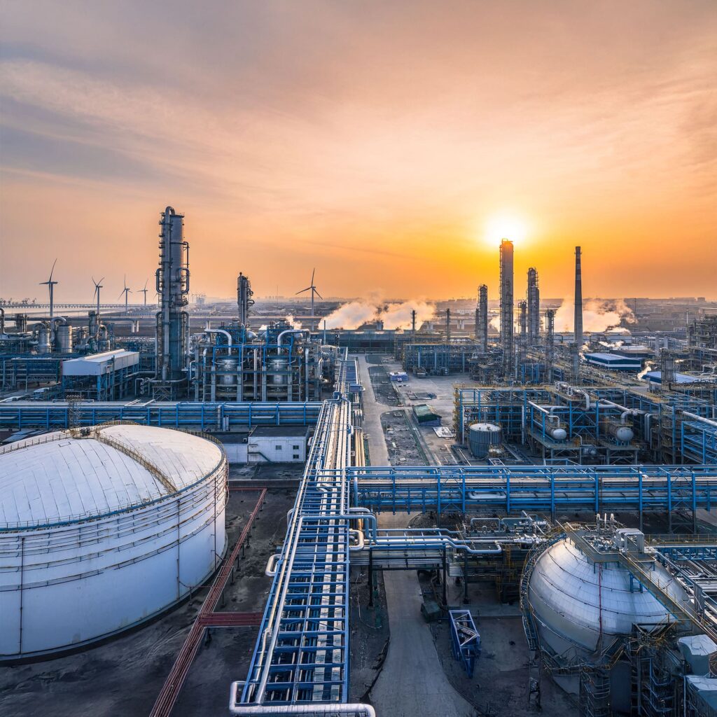 Industrial refinery with storage tanks and tall chimneys at sunrise, emitting steam. Wind turbines are visible in the background under a colorful sky. Numerous pipes run across the complex.