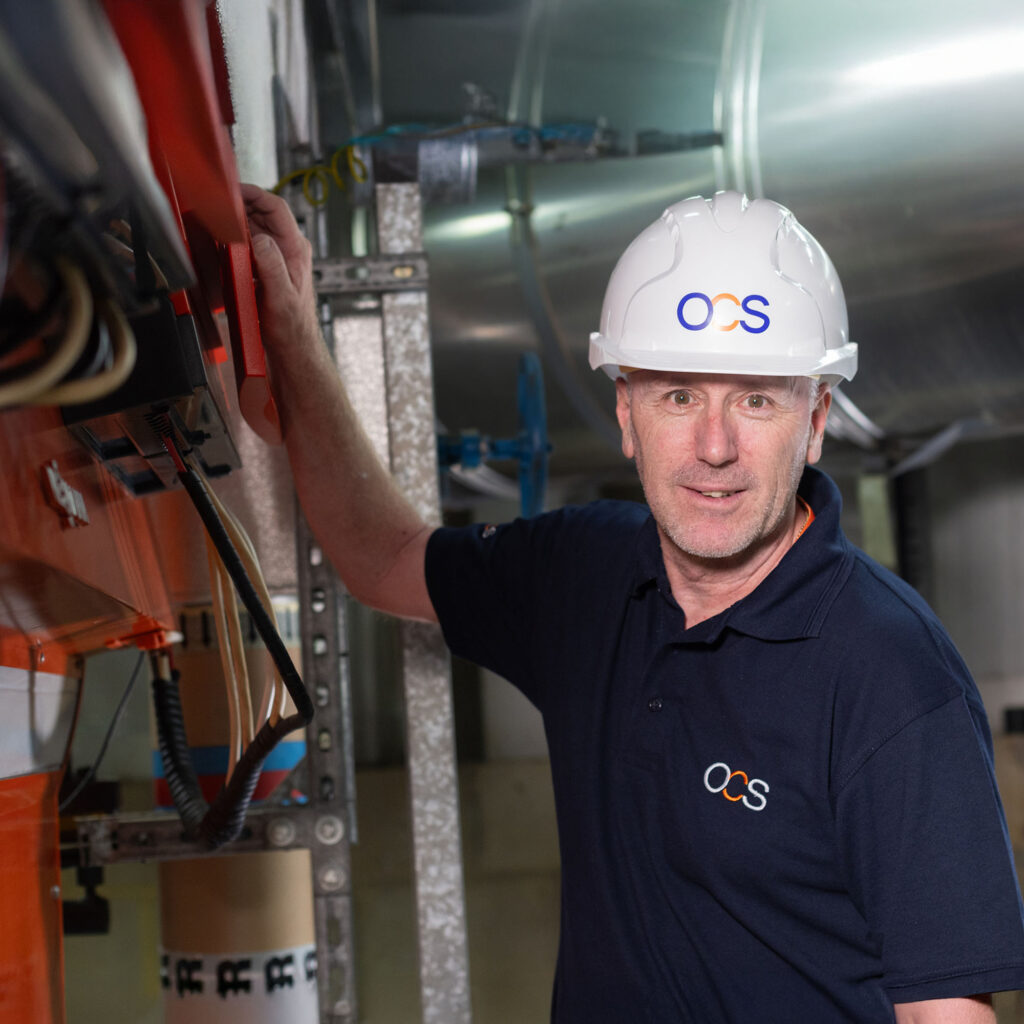 A man wearing a white hard hat and navy blue OCS polo shirt stands in an industrial setting, working with electrical equipment and looking at the camera.