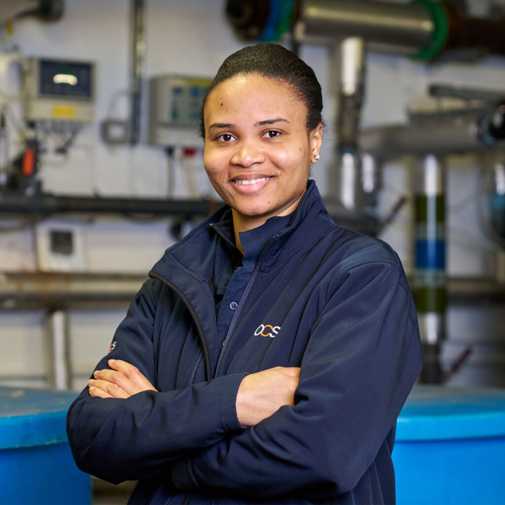 A person wearing a dark jacket stands indoors with arms crossed, smiling at the camera. The background includes industrial equipment and pipes, suggesting a technical or engineering environment.