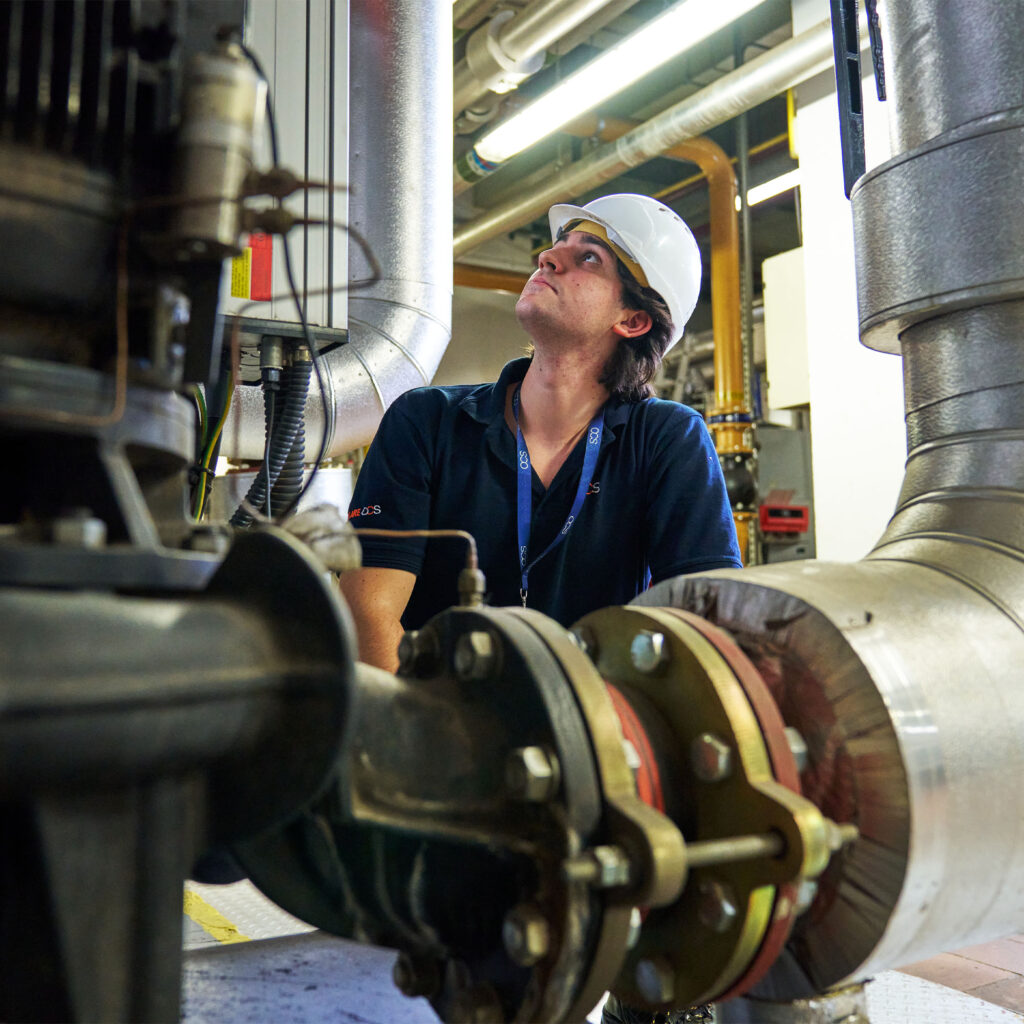 A worker wearing a white hard hat and dark shirt examines large industrial pipes and machinery in a facility with visible metal ducts and equipment.
