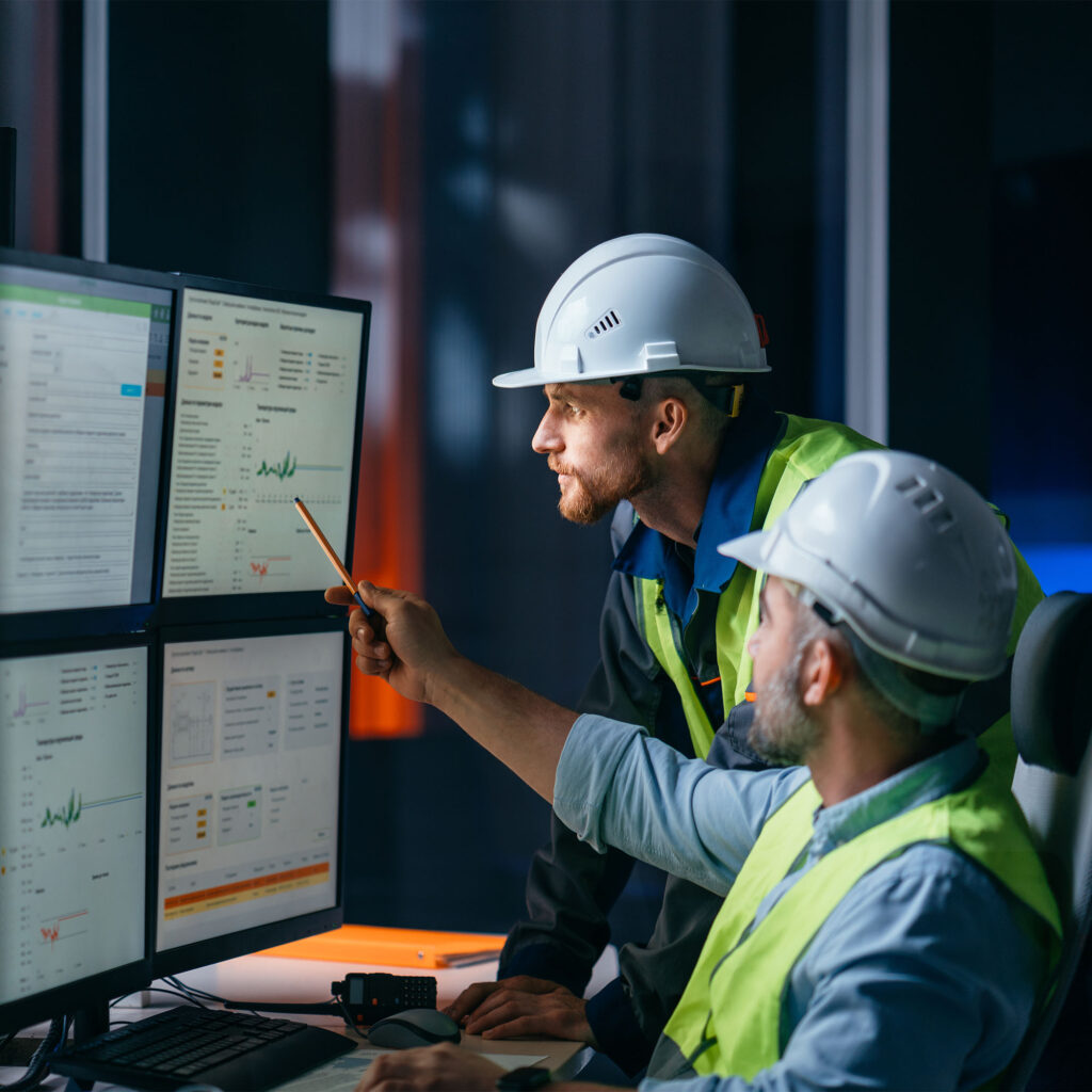 Two engineers wearing safety helmets and vests review data on multiple computer monitors in a control room. One points at a screen displaying charts and graphs while discussing information with the other.