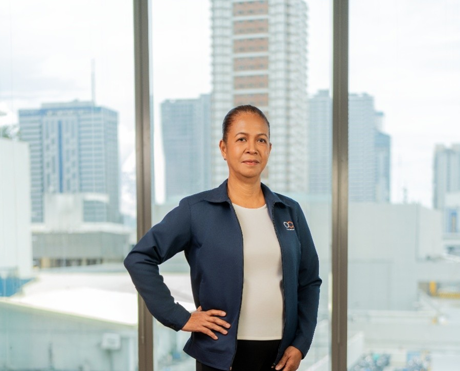 A woman stands indoors in front of large windows with city buildings in the background. She is wearing a navy jacket over a white shirt and has one hand on her hip, looking confidently at the camera.