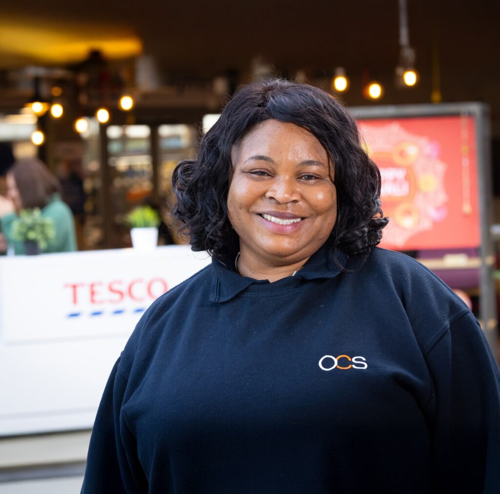 A woman with curly black hair, wearing a black OCS sweatshirt, smiles at the camera. Behind her is a Tesco sign and a brightly lit indoor setting.