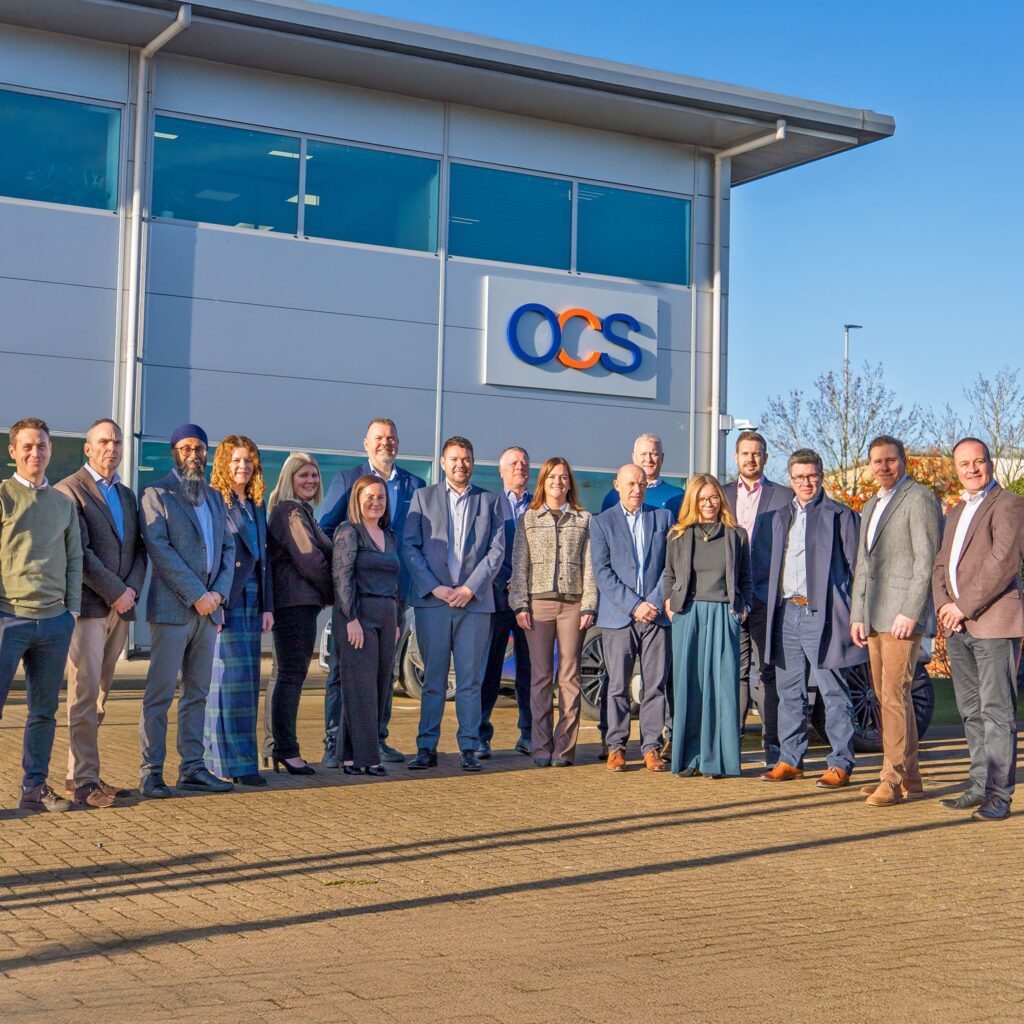 A group of people in business attire stand outside a modern building with an “OCS” sign, posing for a group photo on a sunny day.