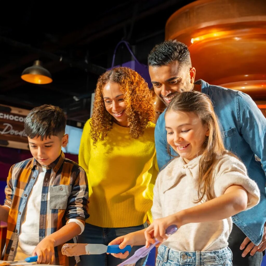 A smiling family of four, two adults and two children, stand together indoors, slicing chocolate treats at a brightly lit counter. The children are focused on cutting while the adults watch and smile behind them.