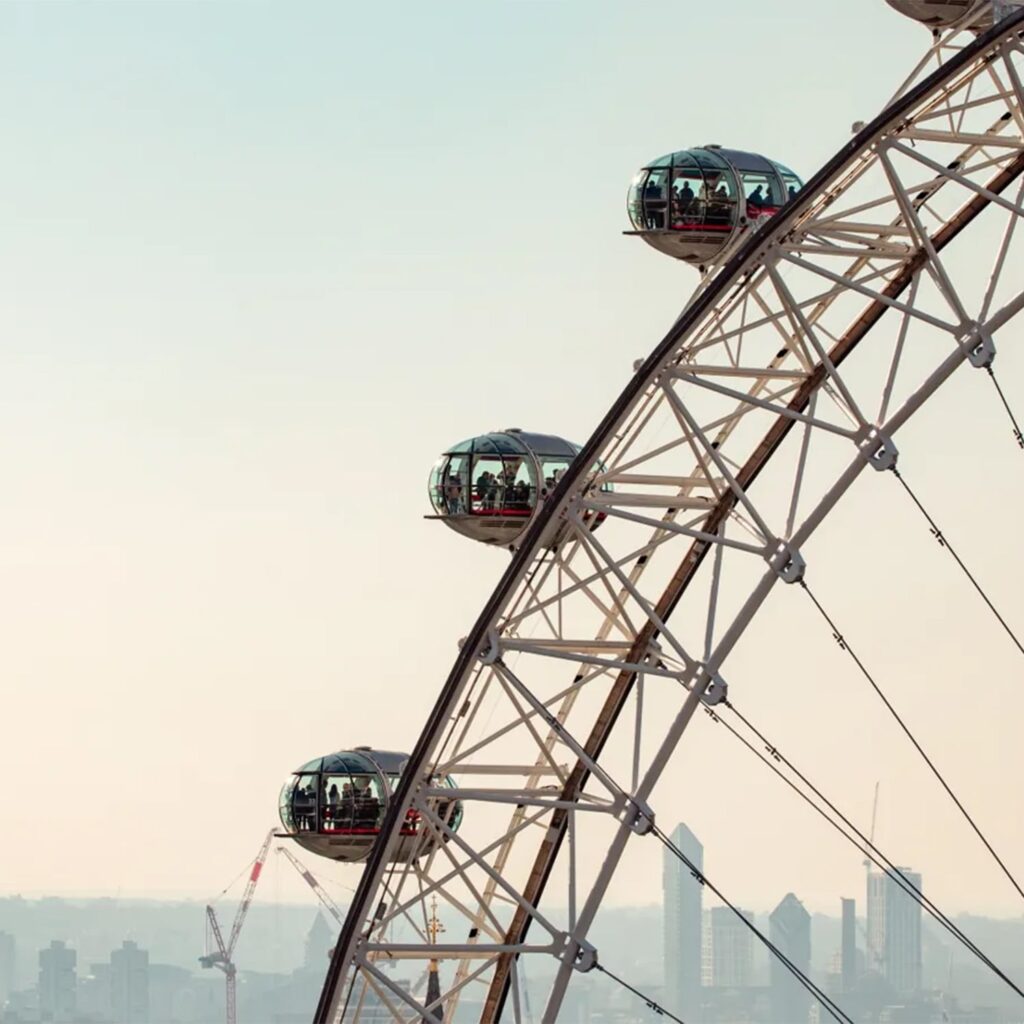 Close-up of four glass pods on a Ferris wheel, likely the London Eye, with a hazy city skyline and cranes visible in the distant background under a pale sky.