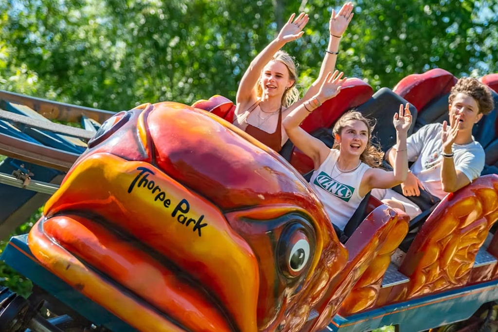 Three young people ride a colorful roller coaster with a fish-shaped front at Thorpe Park, hands in the air and smiling, surrounded by green trees in the background.