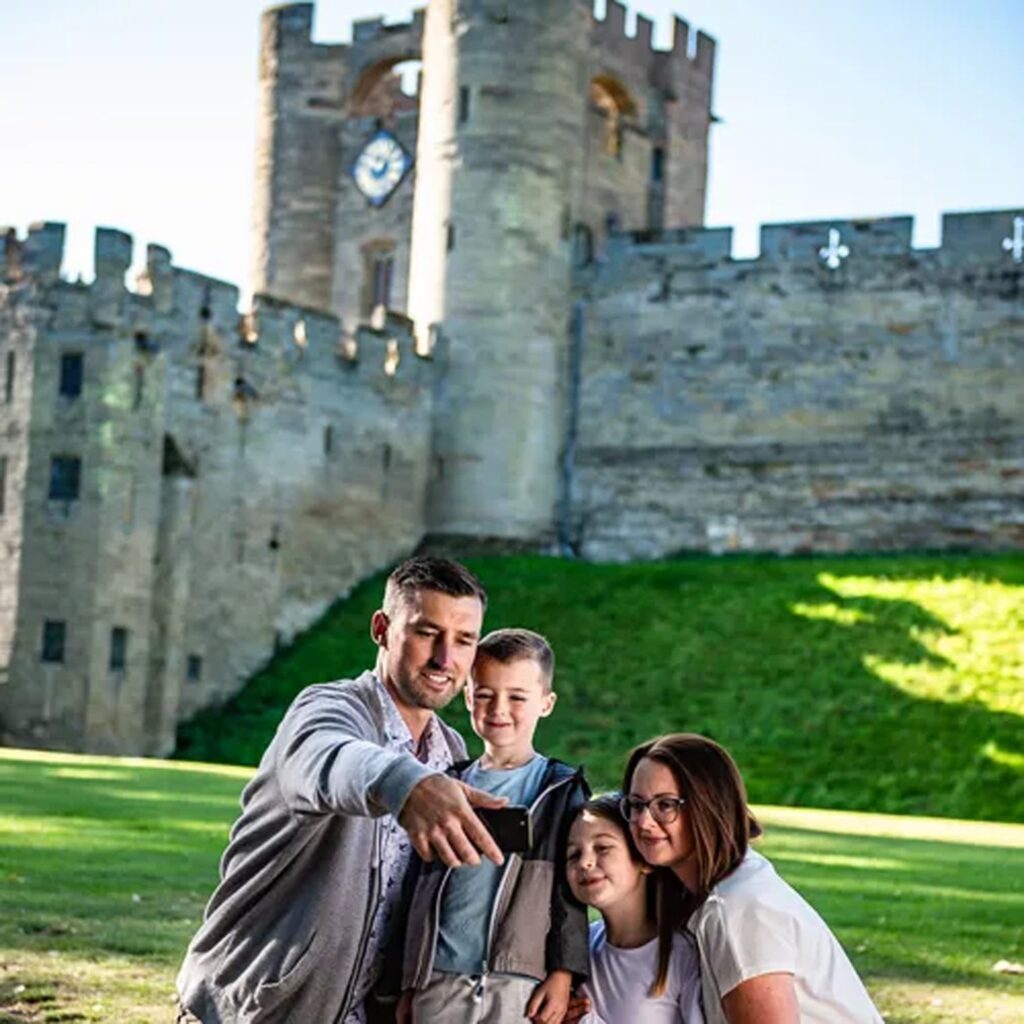 A family of four poses for a selfie in front of a historic stone castle with a clock tower and crenellated walls on a sunny day.