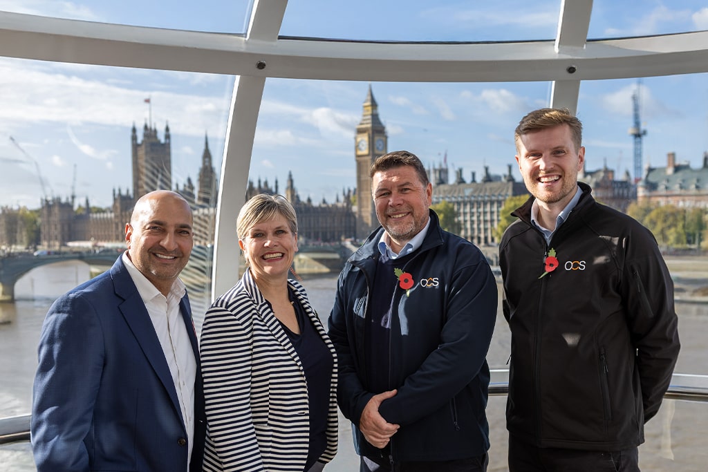 Four people smiling and standing inside a glass capsule with the Houses of Parliament and Big Ben visible in the background; two men wear jackets with OCS logos, all wear poppy pins.