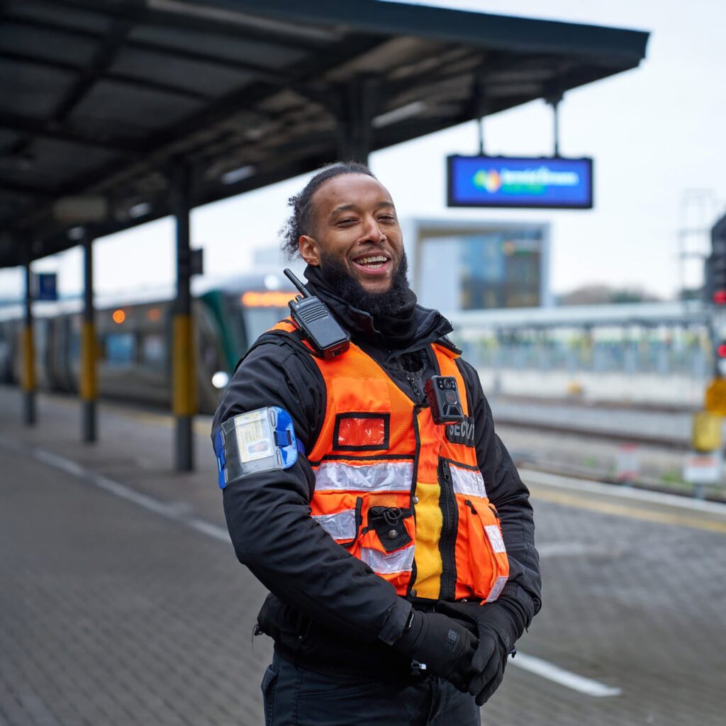 A smiling security officer wearing an orange safety vest and radio stands on a train station platform, with a train and electronic sign in the background.