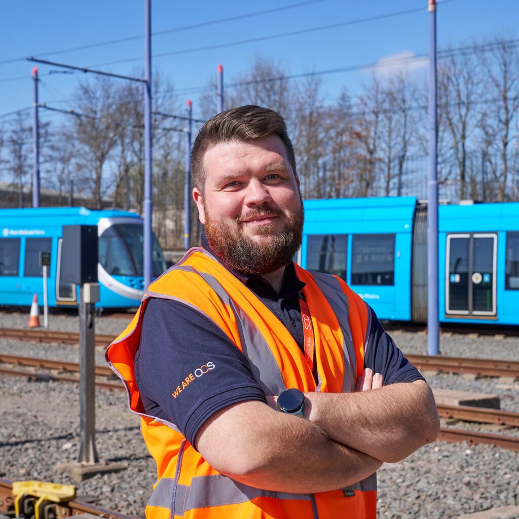 A man in an orange safety vest stands with arms crossed on railway tracks, with blue trams and leafless trees in the background on a sunny day.