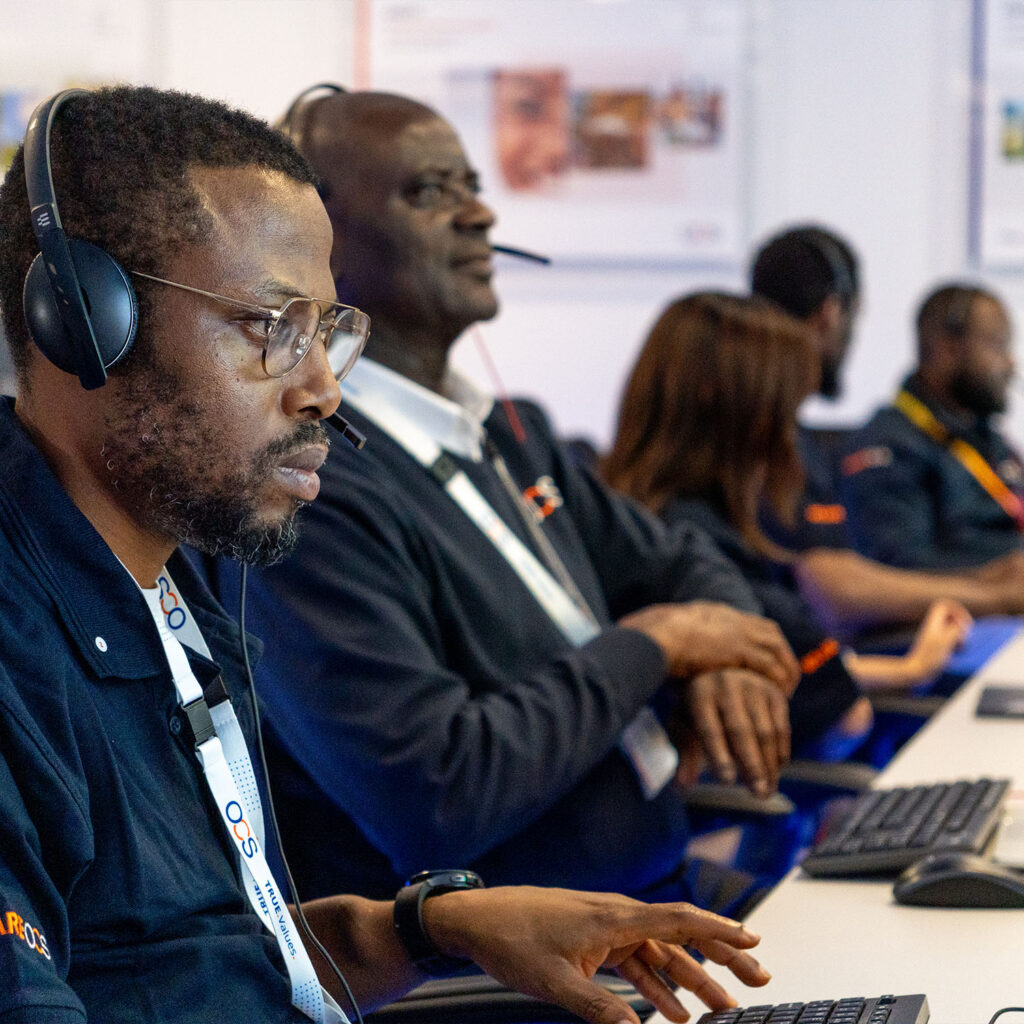 Several people wearing headsets and uniforms sit in a row at desks, focused on computer screens in a modern office or control room environment.