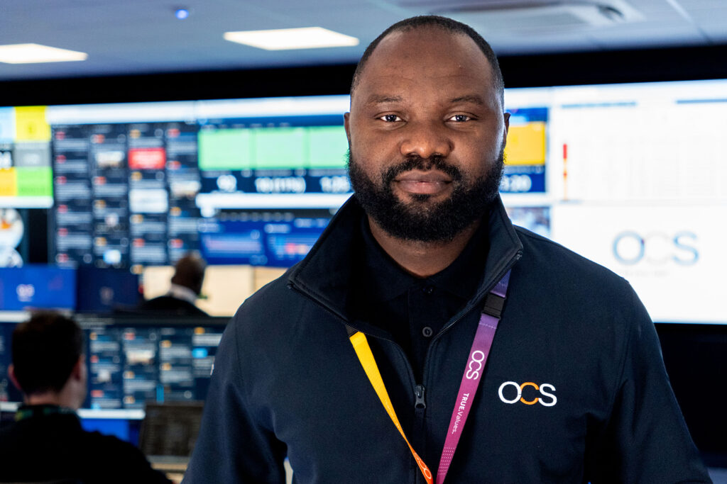 A man wearing an OCS-branded jacket and lanyard stands in a control room with multiple large screens displaying data on the wall behind him. Other people are working at computer stations in the background.