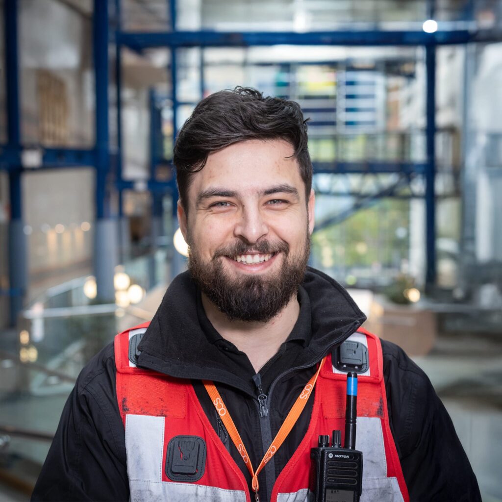 A bearded man wearing a red and white safety vest, black jacket, and lanyard smiles at the camera in an industrial or office setting with blue railings and glass walls in the background.