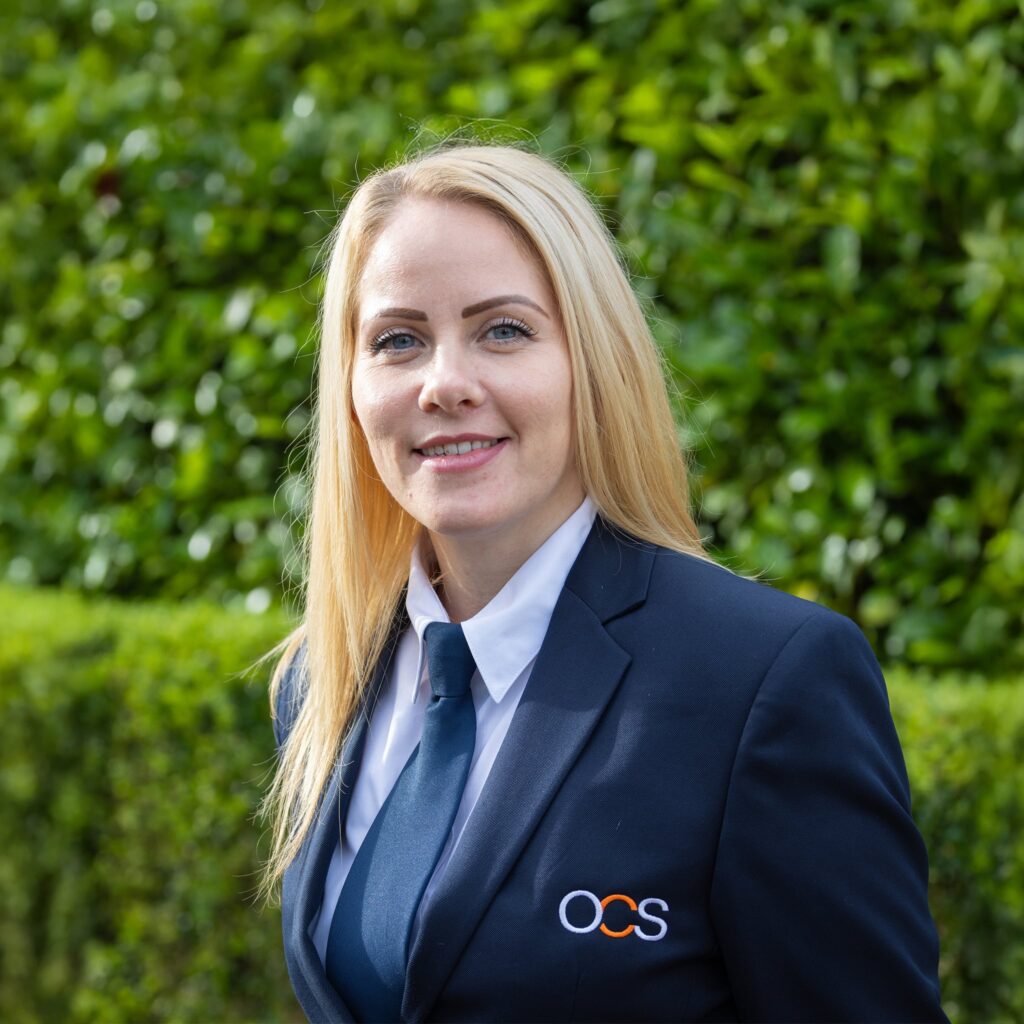 A woman with long blonde hair wearing a navy blue OCS uniform and tie stands outdoors, smiling, with green foliage in the background.