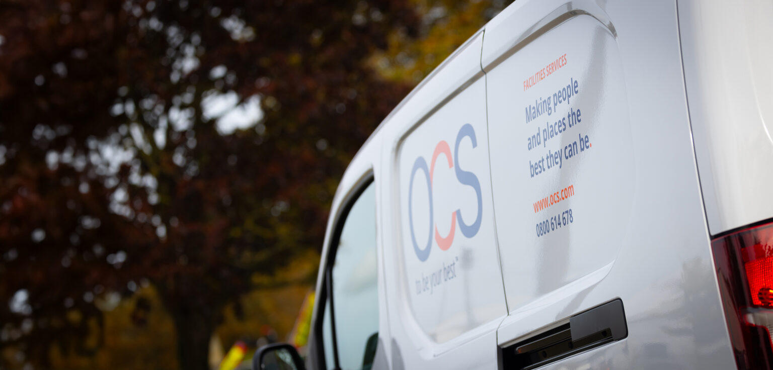 A white van with the OCS logo and slogan, Making people and places the best they can be, parked outdoors next to trees with autumn foliage.