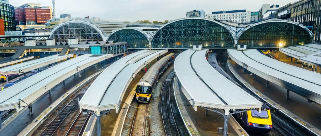 A large train station with multiple covered platforms and two trains, one yellow and black, under an arched glass roof, surrounded by city buildings on a cloudy day.
