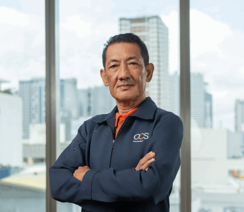 A middle-aged man with short dark hair stands confidently with arms crossed, wearing a navy jacket with OCS logo. He is indoors with large windows showing a cityscape and tall buildings in the background.