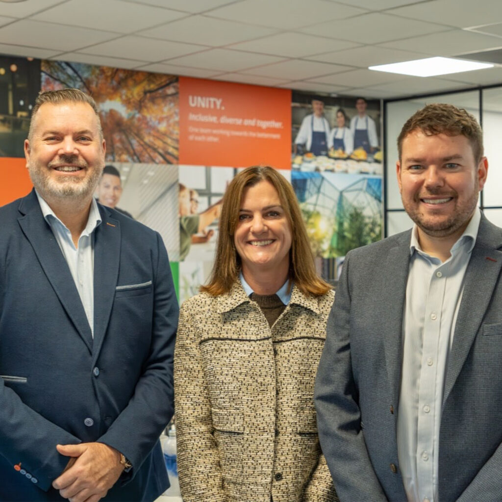 Three people in business attire stand smiling indoors in an office setting. Behind them are posters and images on the wall, including one with the word UNITY. The space is well-lit with a modern design.