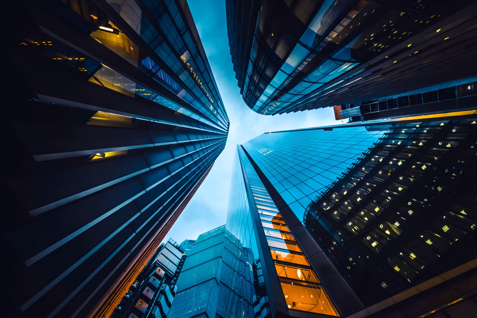 Dramatic upward view of tall, modern skyscrapers with reflective glass surfaces, illuminated windows, and a blue sky peeking through the narrow gap between the buildings.