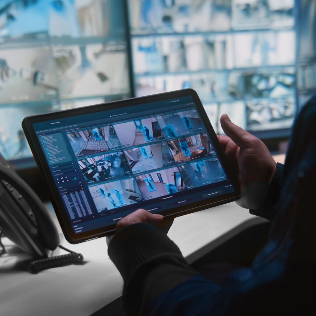 A person holds a tablet displaying multiple security camera feeds, while a wall of surveillance monitors shows similar footage in the background.