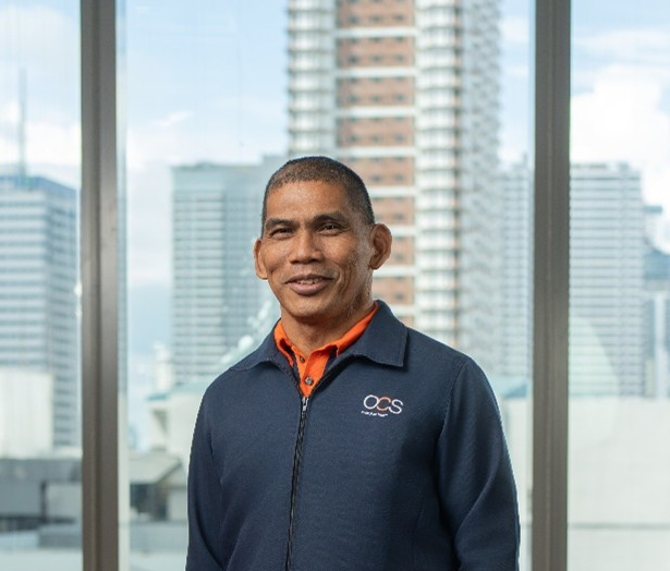 A man in a navy OCS jacket and orange shirt stands indoors, smiling, with tall city buildings visible through large windows behind him.