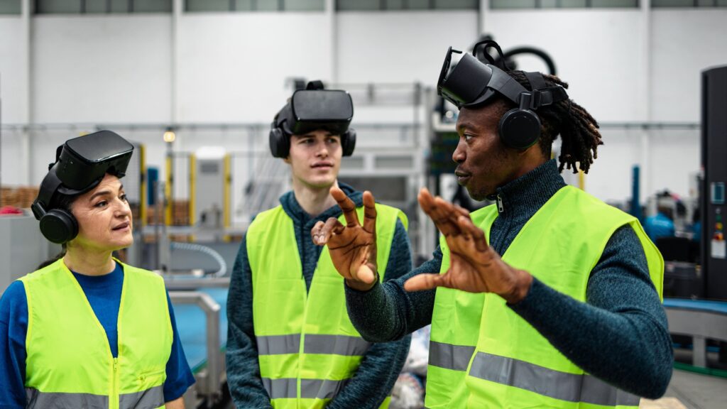 Three people wearing VR headsets and high-visibility vests stand in a factory, engaged in discussion. One person gestures with their hands while the others listen attentively. Industrial equipment is visible in the background.