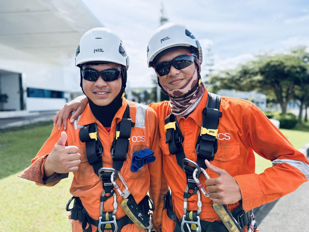 Two workers in orange safety uniforms, helmets, sunglasses, and harnesses stand outdoors, smiling at the camera. One gives a thumbs up while the other holds a tool. They appear to be at an industrial or construction site.