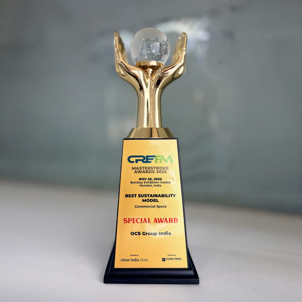 A gold and black trophy with a figure holding a glass globe on top. The plaque reads: CRE FM Masterstroke Award 2023, Best Sustainability Model, Special Award, OCS Group India, with event details and logos.