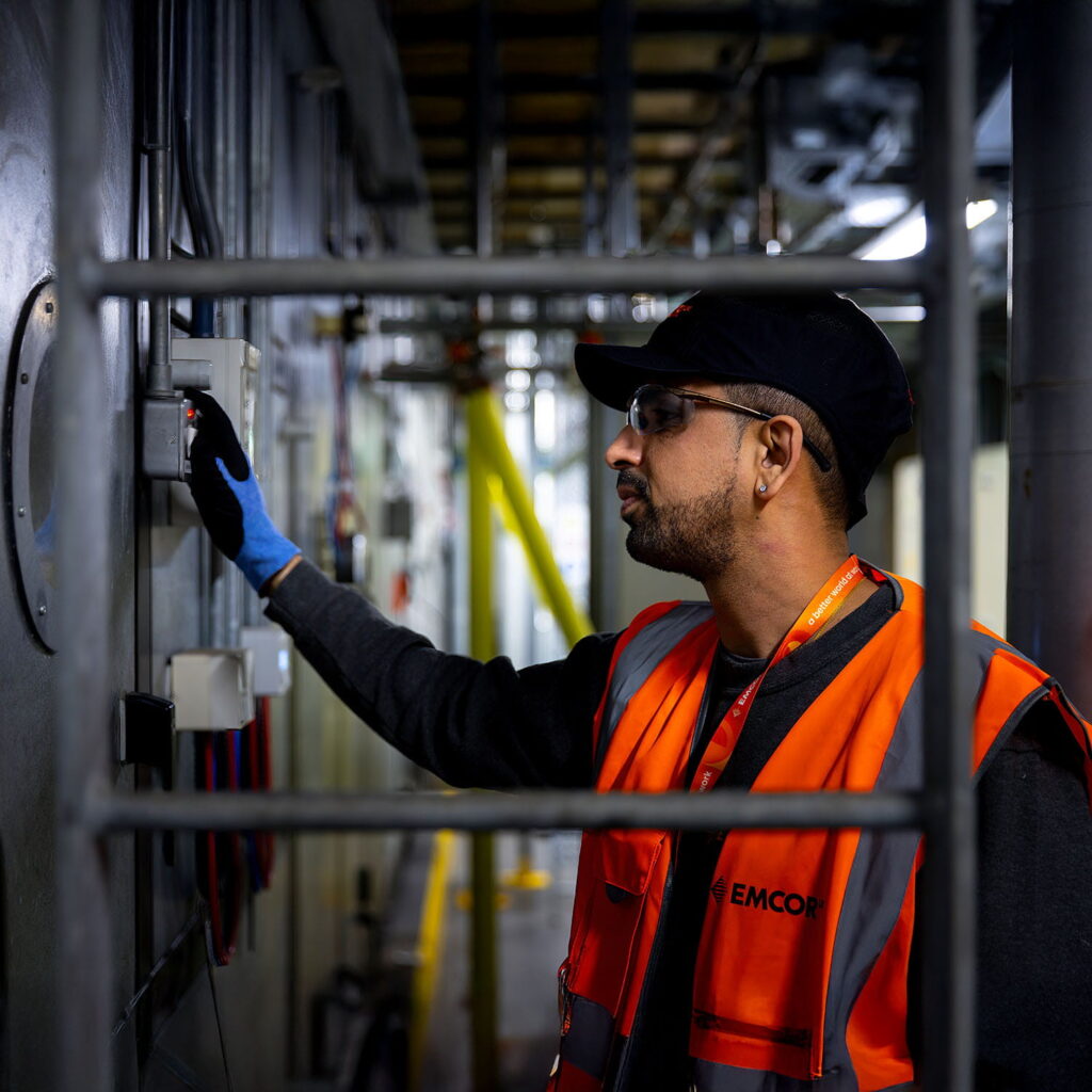A man wearing an orange safety vest, cap, and blue gloves operates a control panel in an industrial setting, surrounded by metal pipes and machinery.