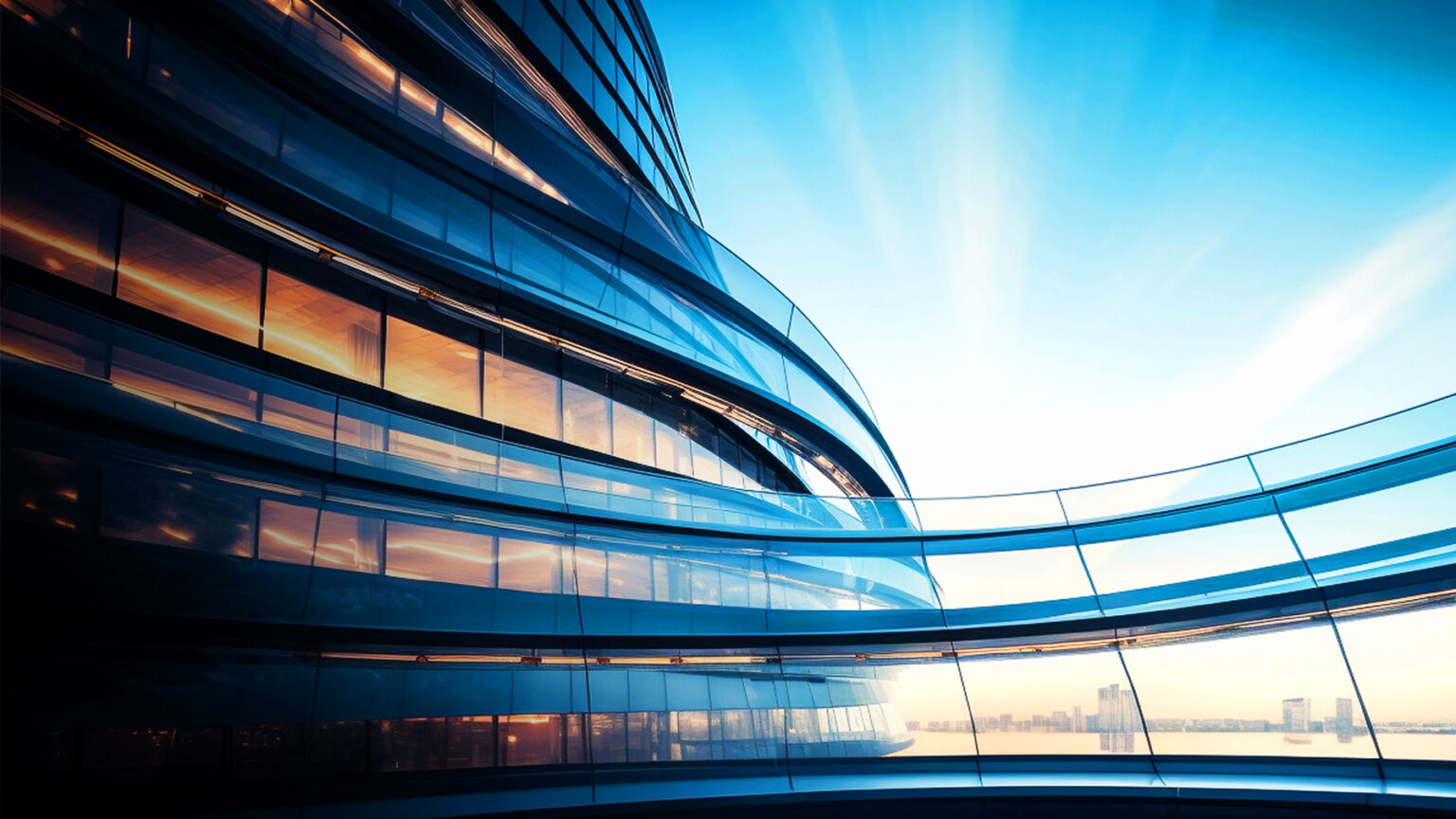 Curved glass exterior of a modern building reflects sunlight at sunrise or sunset, with views of a city skyline and water in the background under a clear blue sky.