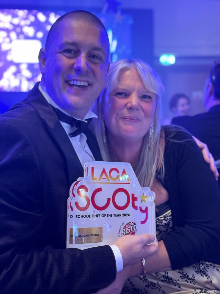A smiling man in a tuxedo holds a trophy reading LACA School Chef of the Year 2024 while posing with a smiling woman at an indoor awards event. The background is lit with blue and purple lights.