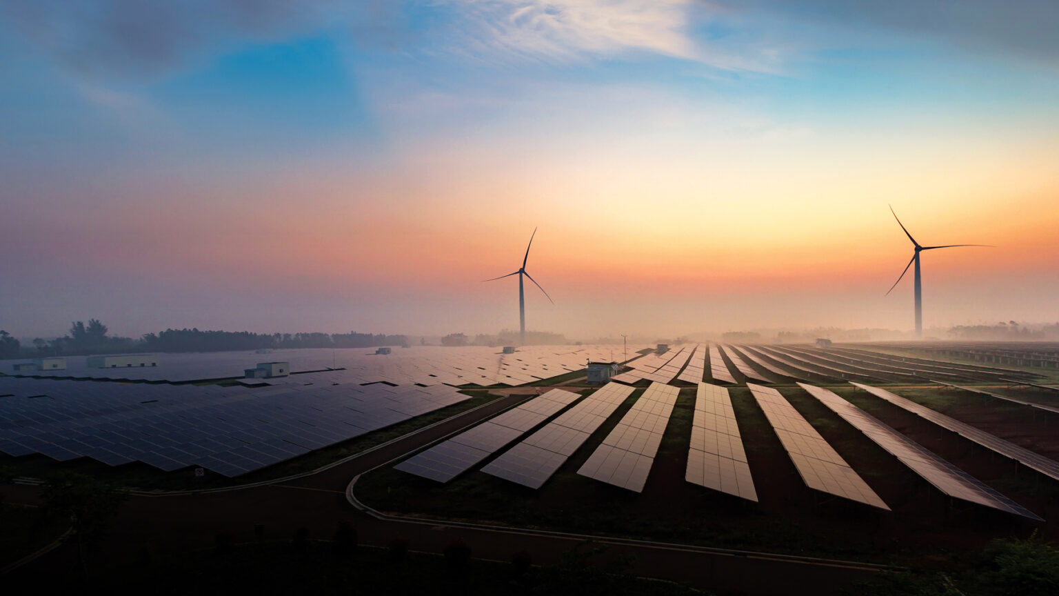 Rows of solar panels and two large wind turbines sit in a field at sunrise or sunset, with a colorful sky and mist in the distance, illustrating renewable energy sources in a modern landscape.