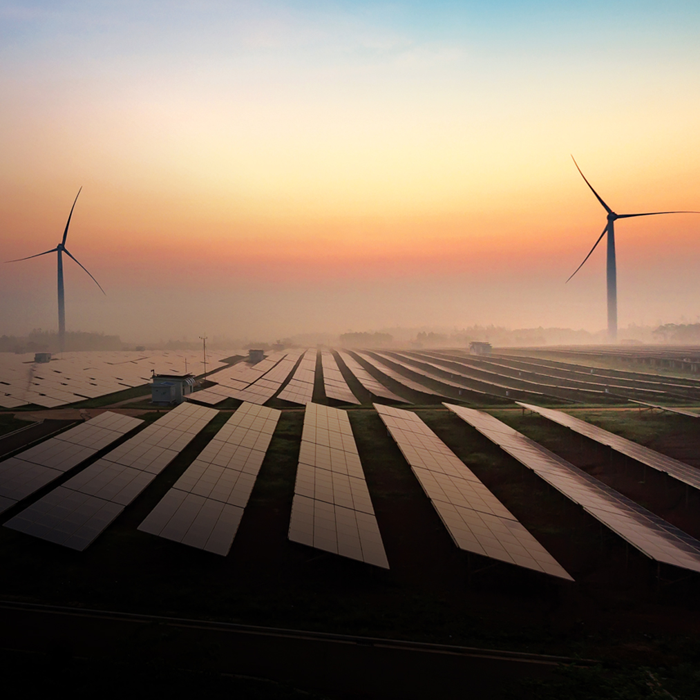 Rows of solar panels and two wind turbines are seen in a field during sunrise or sunset, with a colorful sky and mist on the horizon, symbolizing renewable energy and sustainability.