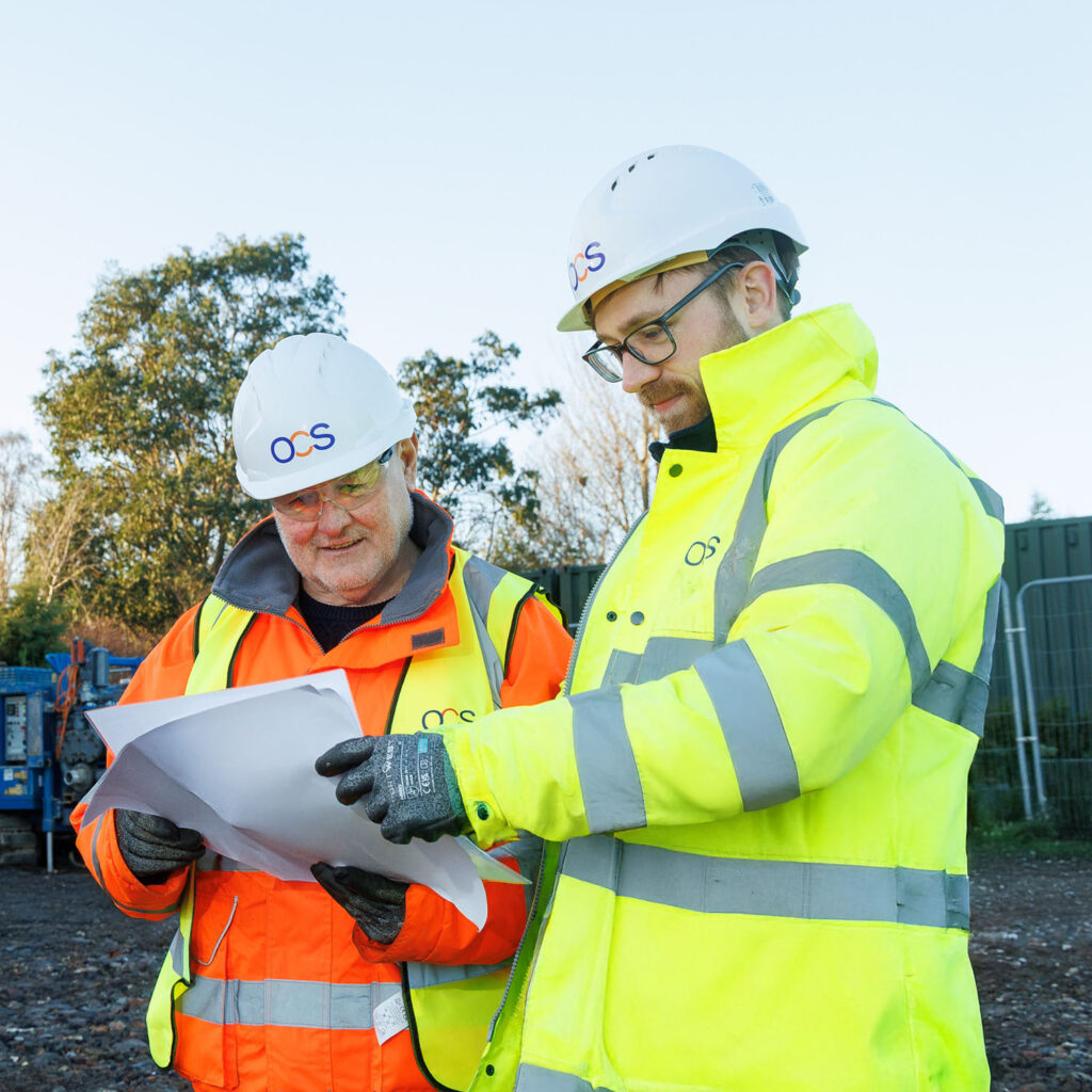Two construction workers in high-visibility jackets and OCS-branded hard hats review plans together outdoors at a worksite, with trees and equipment visible in the background.