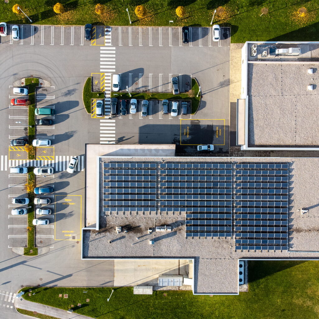 Aerial view of a parking lot next to a building with solar panels on the roof, surrounded by green grass and parked cars in marked spaces.