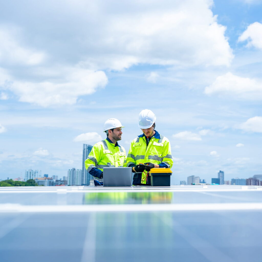 Two engineers in safety gear stand beside a laptop and equipment on a rooftop with solar panels, discussing work. A city skyline and blue sky with clouds are visible in the background.