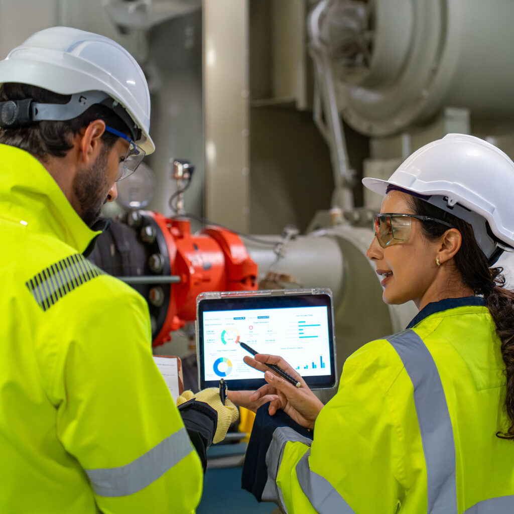 Two engineers in safety gear and helmets examine data on a tablet in an industrial setting, discussing charts and graphs displayed on the screen. Machinery and technical equipment are visible in the background.