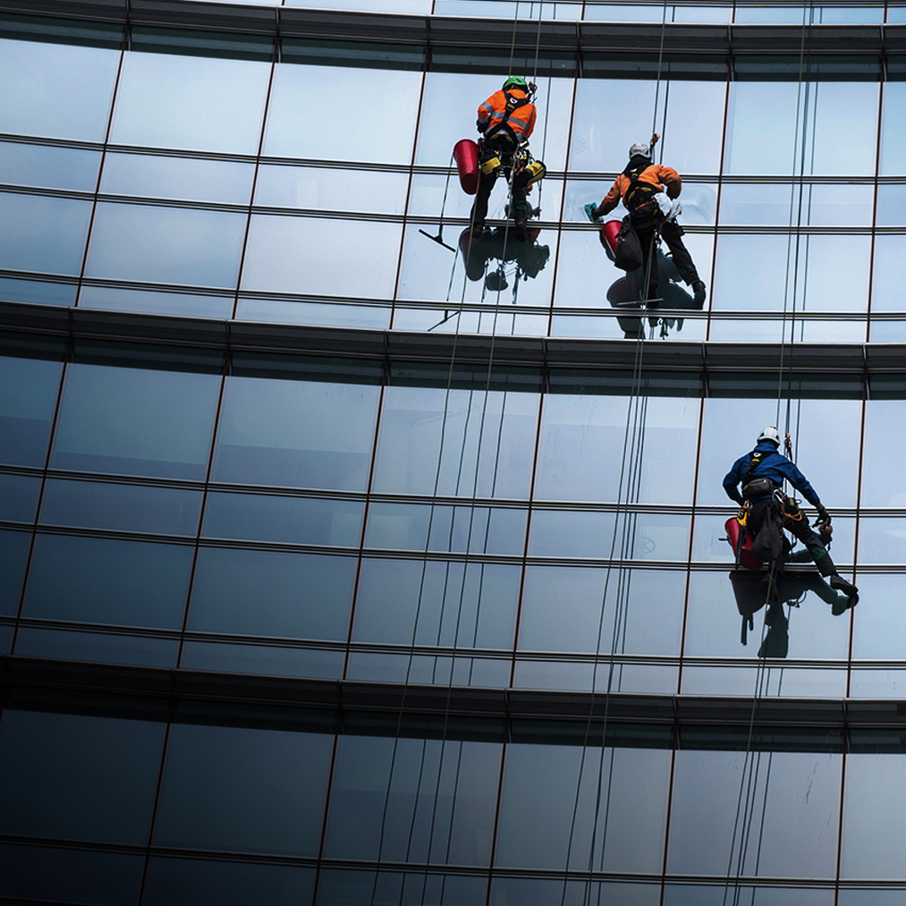 Three workers in safety gear are suspended by ropes, cleaning the exterior glass windows of a tall modern building. Their reflections are visible on the shiny surface of the windows.