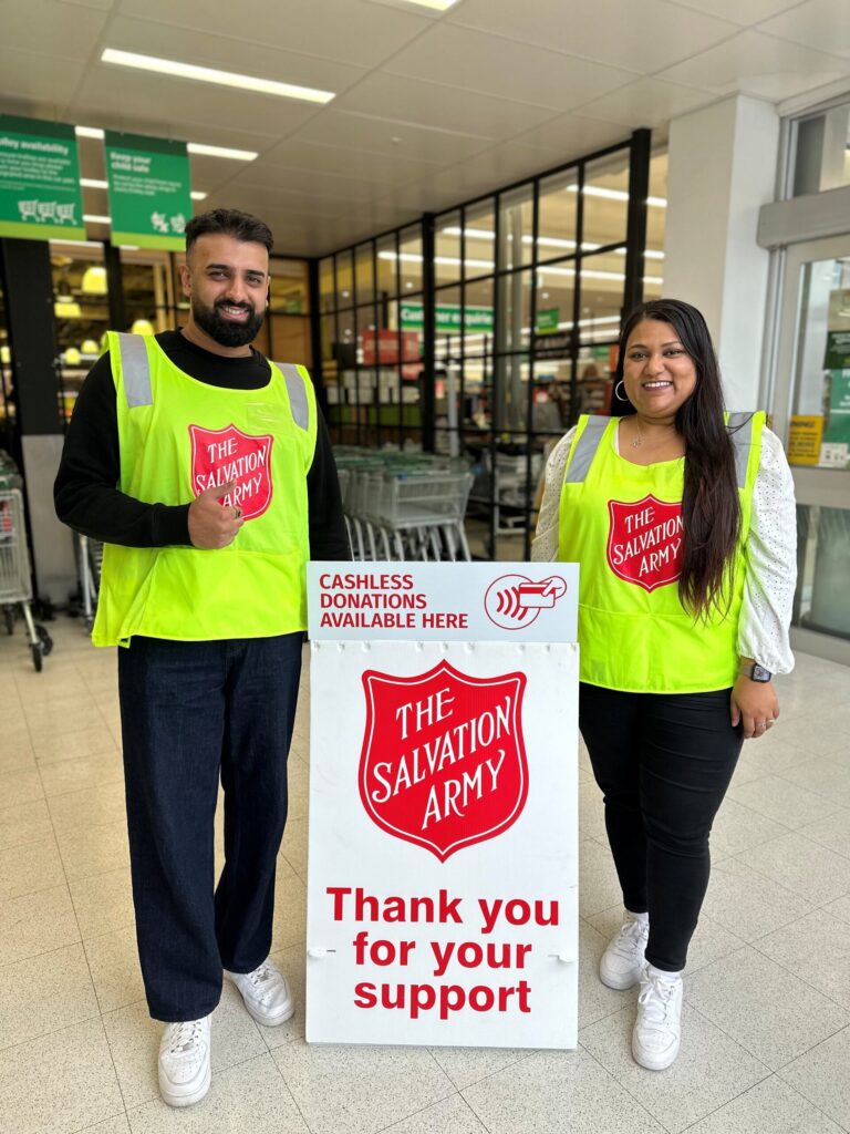 Two volunteers wearing neon yellow vests with Salvation Army logos stand smiling beside a Salvation Army donation sign inside a store entrance. The sign reads Thank you for your support and promotes cashless donations.