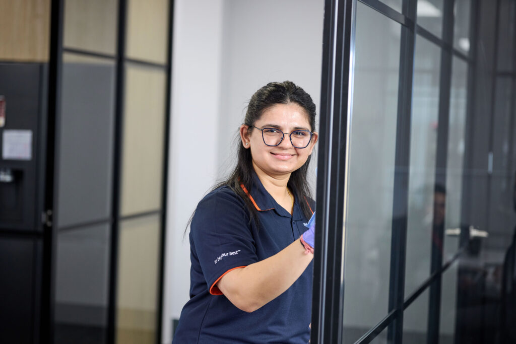 A woman wearing glasses and a navy shirt smiles while cleaning a glass wall in a modern office environment.