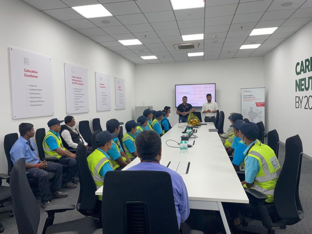 A group of workers in blue uniforms and safety vests sit around a conference table, listening to two men presenting near a screen in a modern meeting room with posters on the walls.