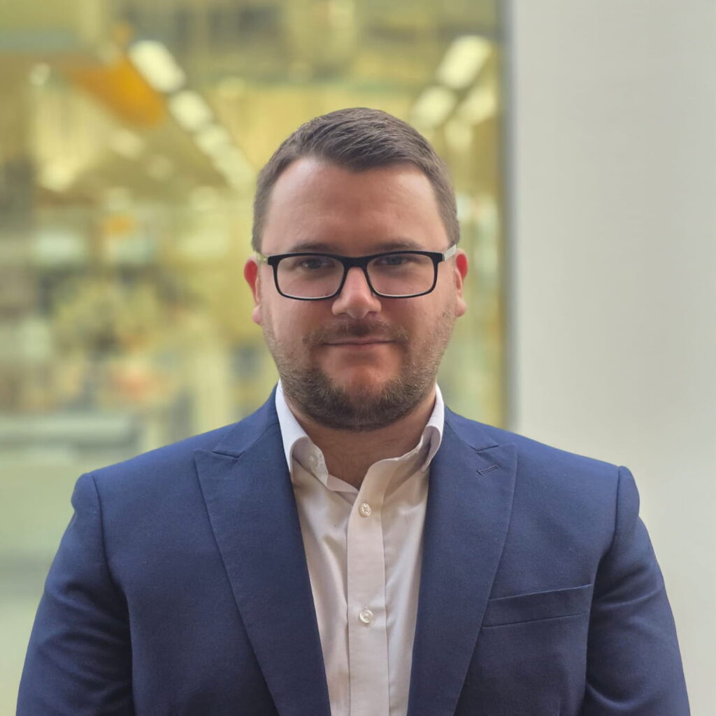 A man with short hair, a trimmed beard, and glasses wears a navy blue suit and white shirt, standing indoors with a blurred background of lights and glass panels.