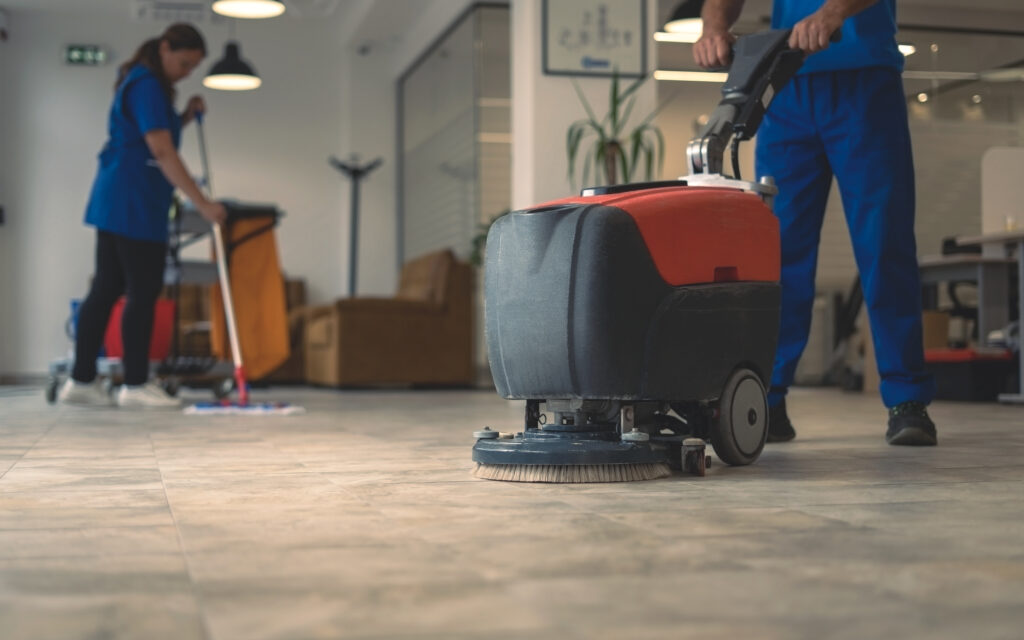 Two people in blue uniforms are cleaning a large indoor floor; one is using a red floor scrubber machine and the other is mopping in the background. The setting appears to be a modern office or commercial space.