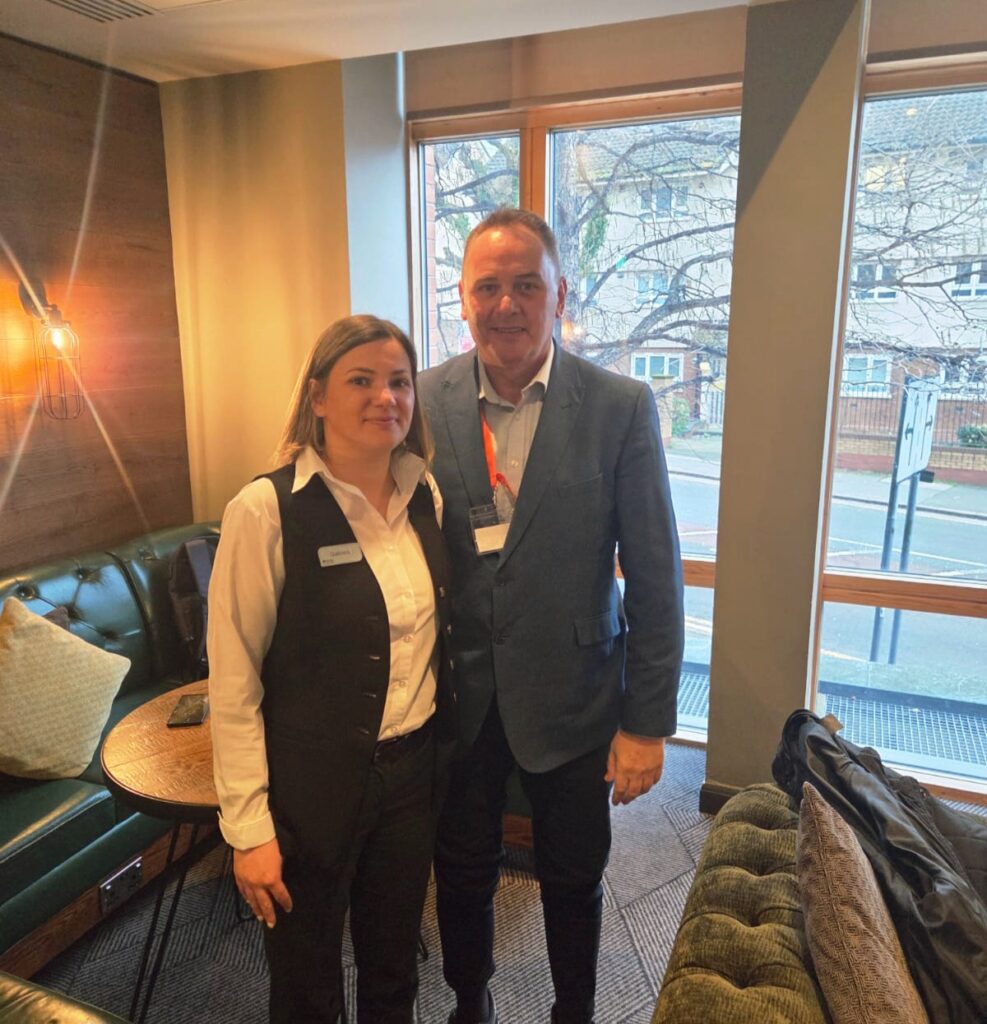 A woman in a white shirt and black vest stands next to a man in a blue blazer. They are indoors, smiling at the camera, with a window and trees visible in the background.
