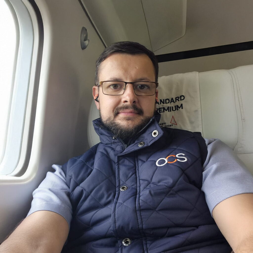 A man with glasses and a trimmed beard sits on an airplane next to a window. He wears a blue quilted vest with an OCS logo over a light blue shirt. The seat cover behind him reads STANDARD PREMIUM.